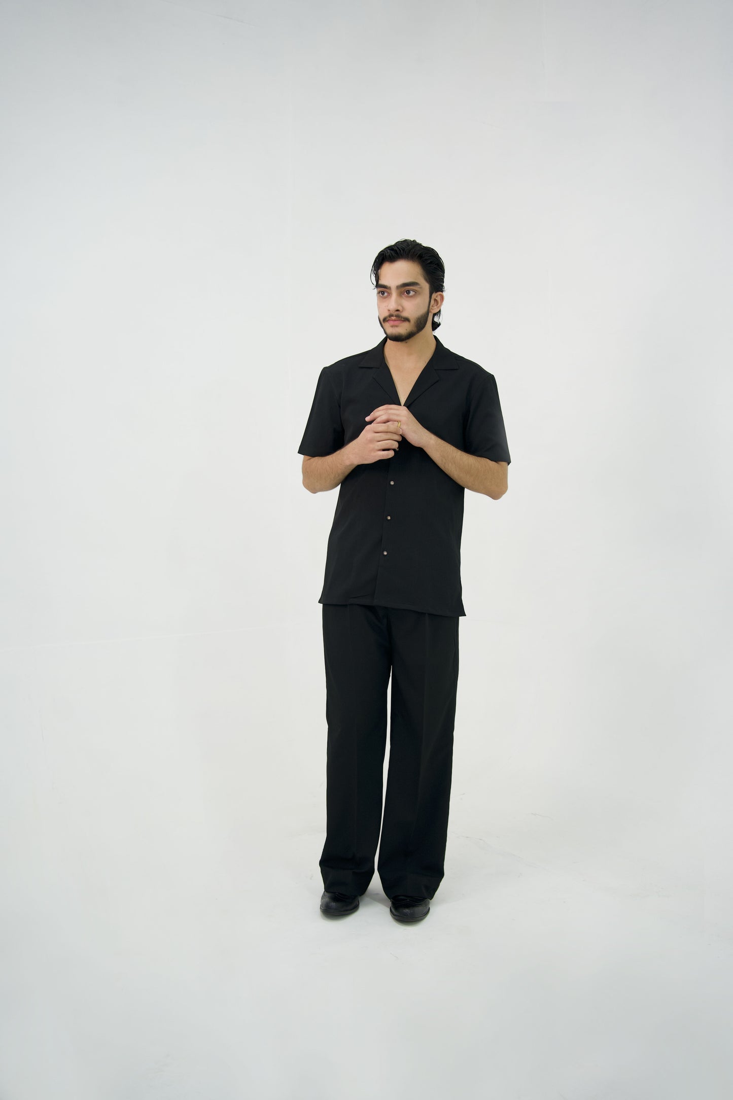 Man posing in a black cuban collar shirt and matching tailored trousers by Abbod Atelier on a white studio background.