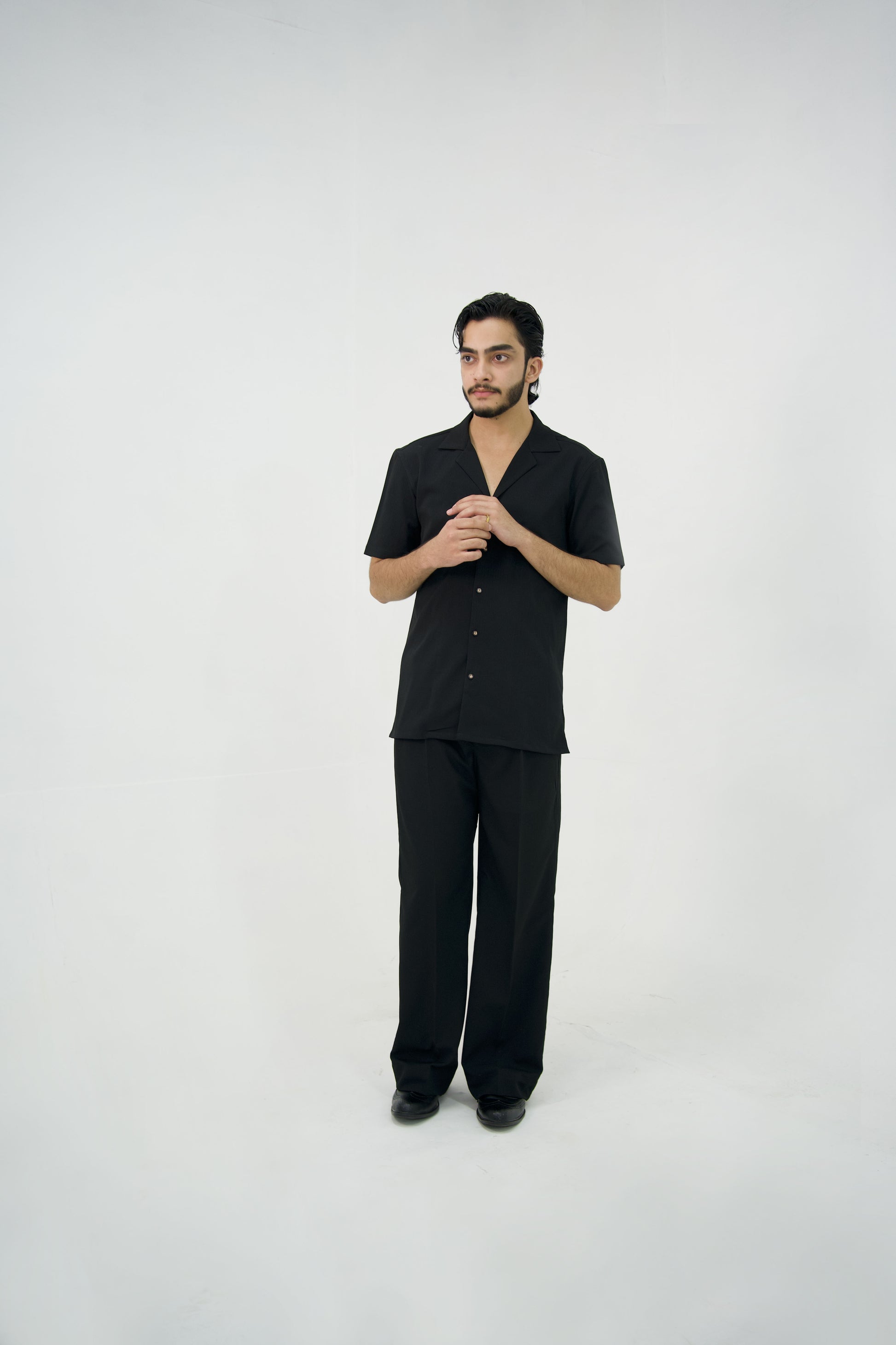 Man posing in a black cuban collar shirt and matching tailored trousers by Abbod Atelier on a white studio background.
