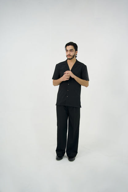 Man posing in a black cuban collar shirt and matching tailored trousers by Abbod Atelier on a white studio background.