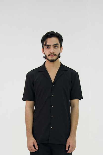 Man posing in a black cuban collar shirt by Abbod Atelier on a white studio background.