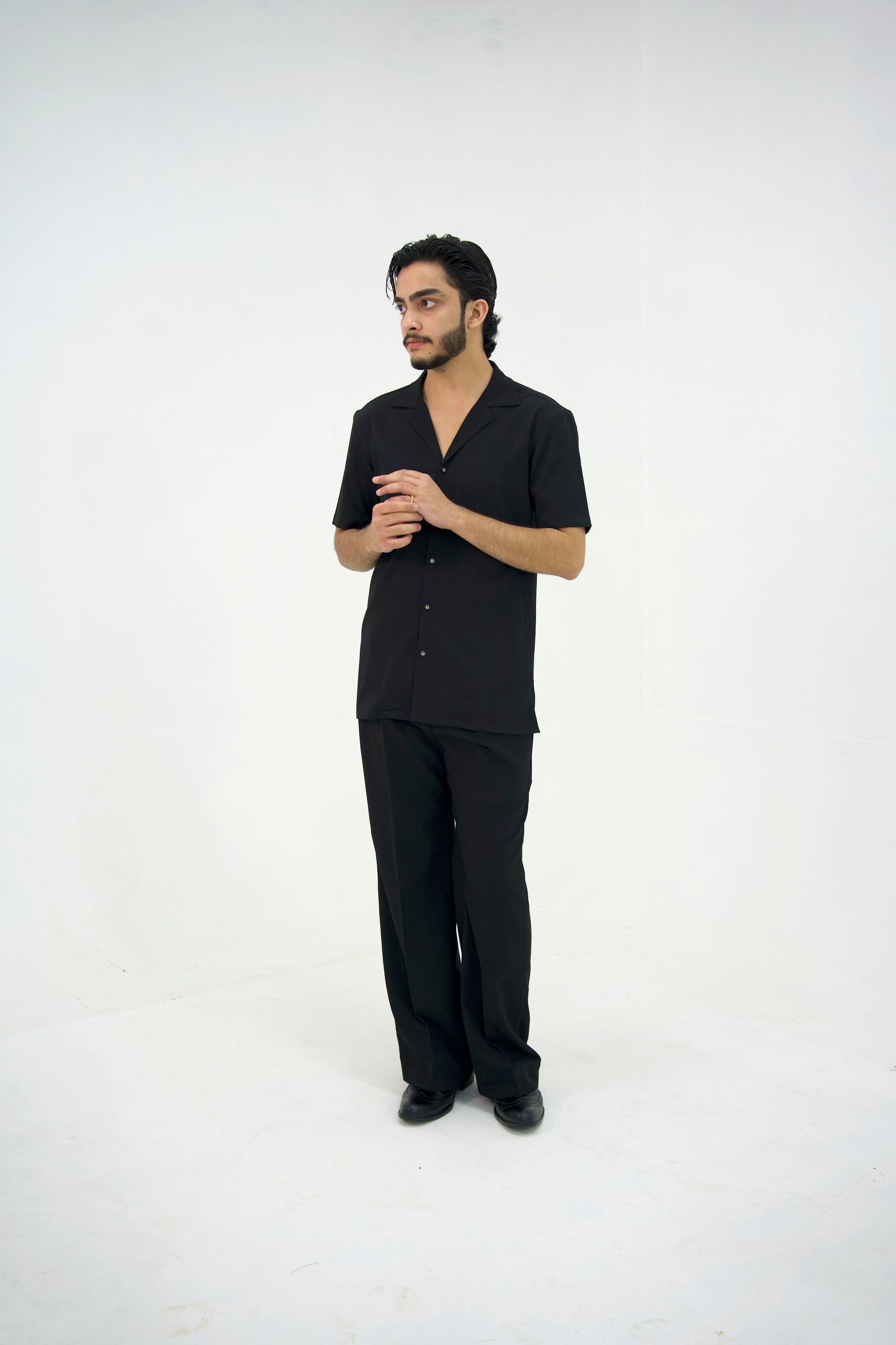 Man posing in a black cuban collar shirt and matching tailored trousers by Abbod Atelier on a white studio background.