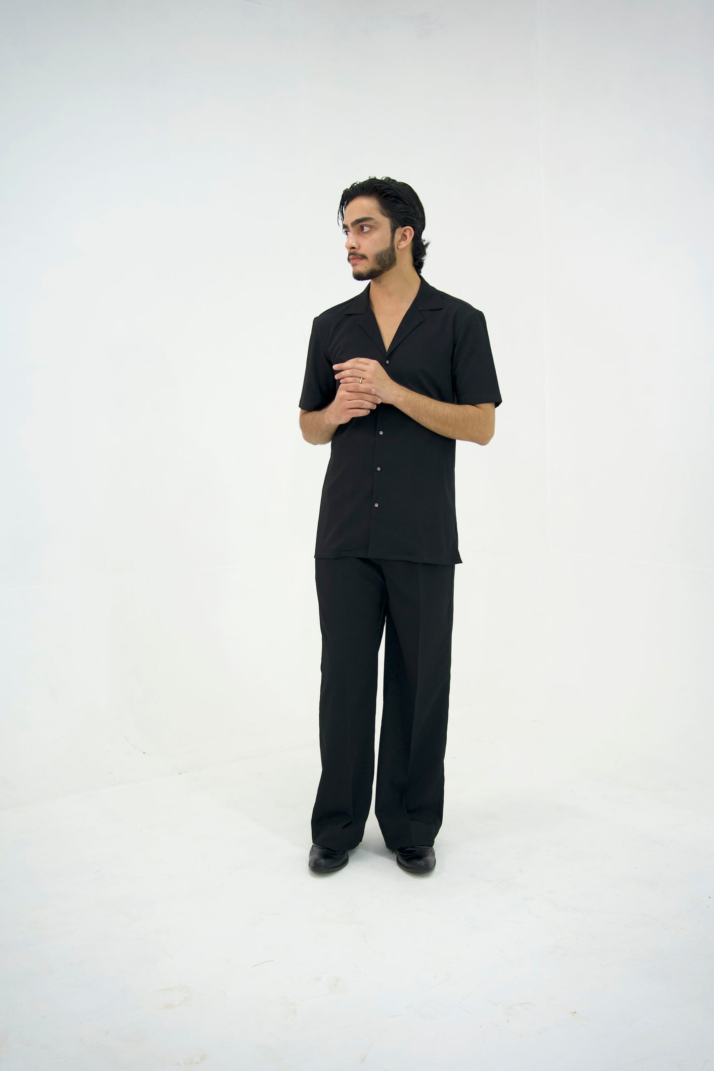 Man posing in a black cuban collar shirt and matching tailored trousers by Abbod Atelier on a white studio background.