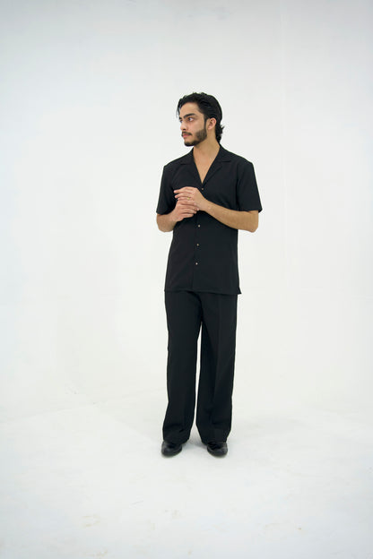 Man posing in a black cuban collar shirt and matching tailored trousers by Abbod Atelier on a white studio background.