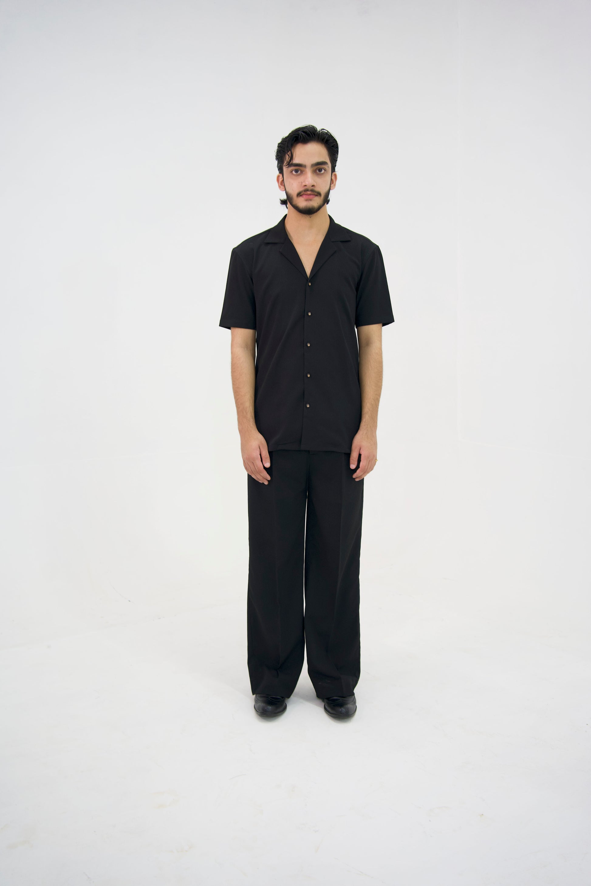 Man posing in a black cuban collar shirt and matching tailored trousers by Abbod Atelier on a white studio background.