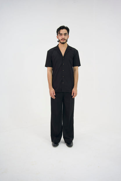 Man posing in a black cuban collar shirt and matching tailored trousers by Abbod Atelier on a white studio background.