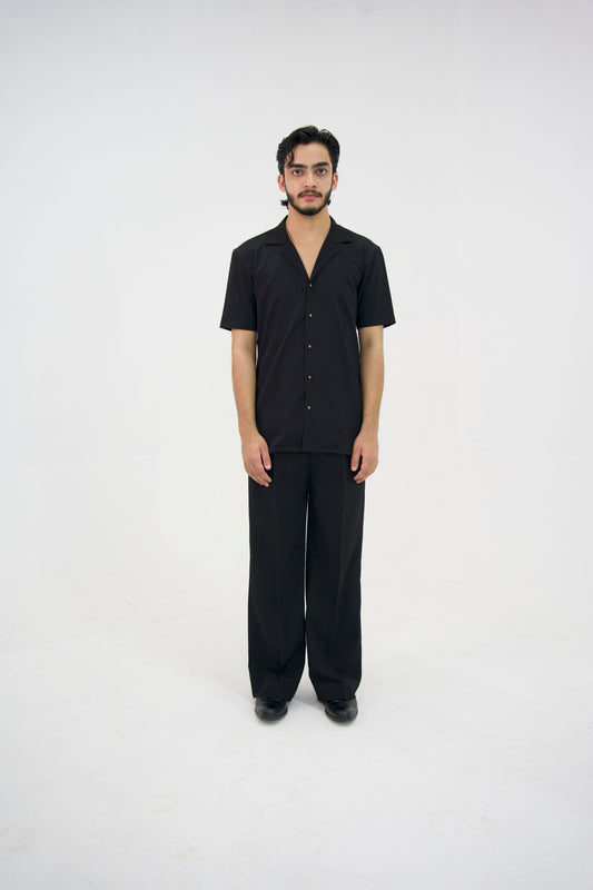 Man posing in a black cuban collar shirt and matching tailored trousers by Abbod Atelier on a white studio background.