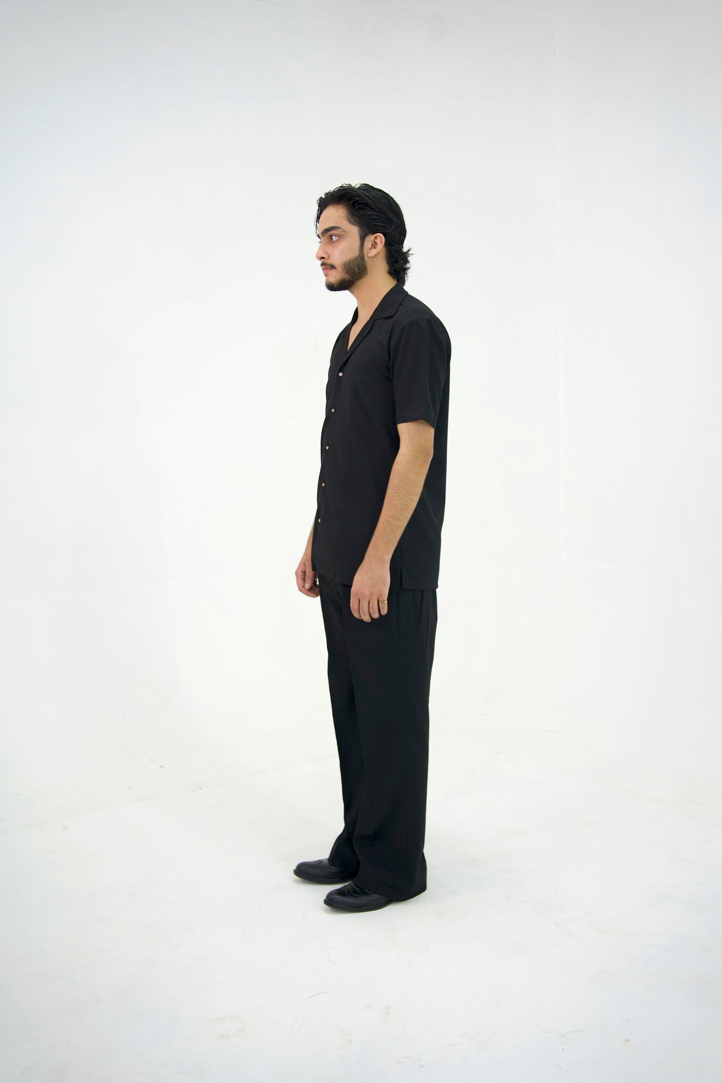 Man posing in a black cuban collar shirt and matching tailored trousers by Abbod Atelier on a white studio background.