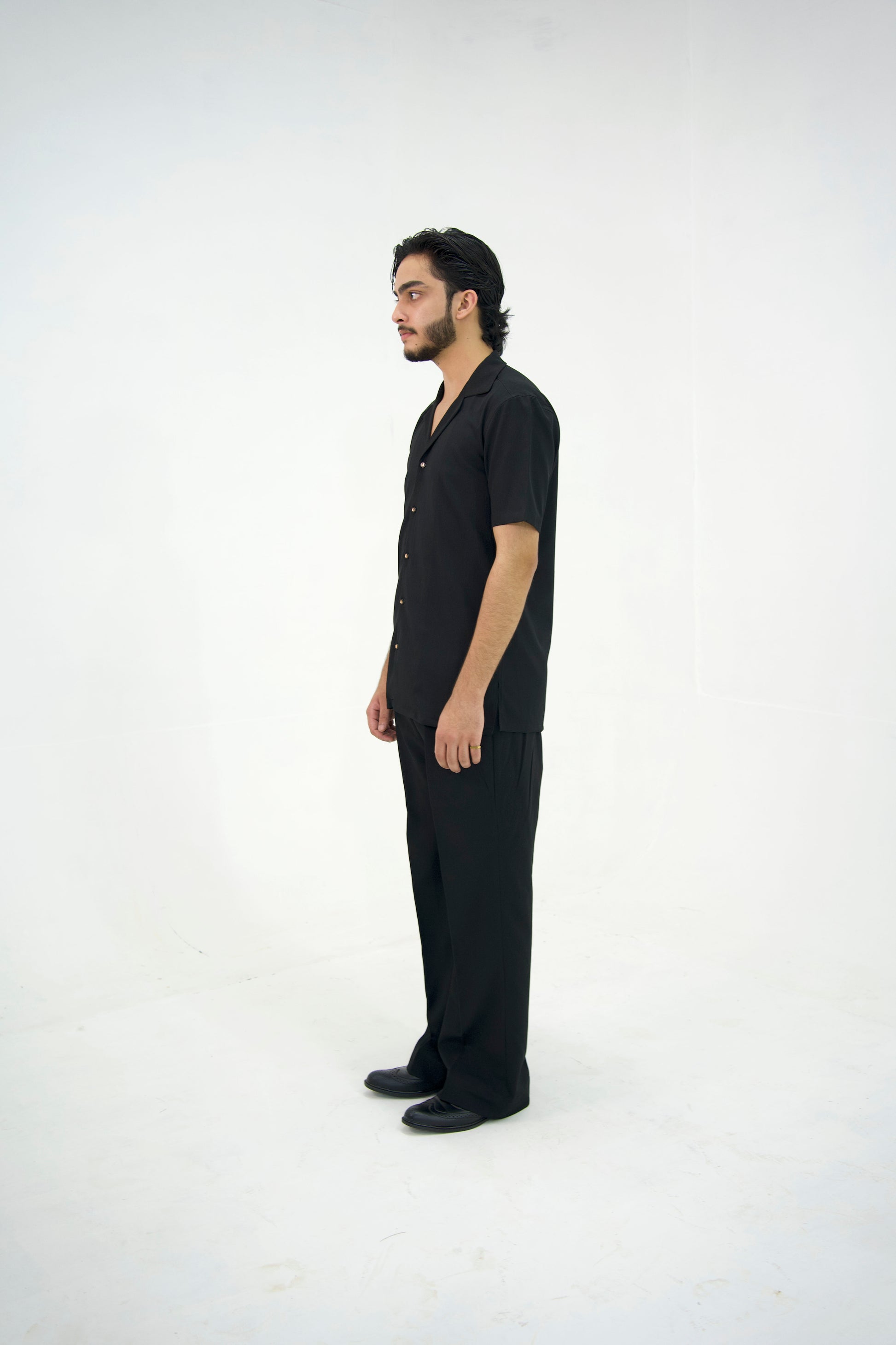 Man posing in a black cuban collar shirt and matching tailored trousers by Abbod Atelier on a white studio background.