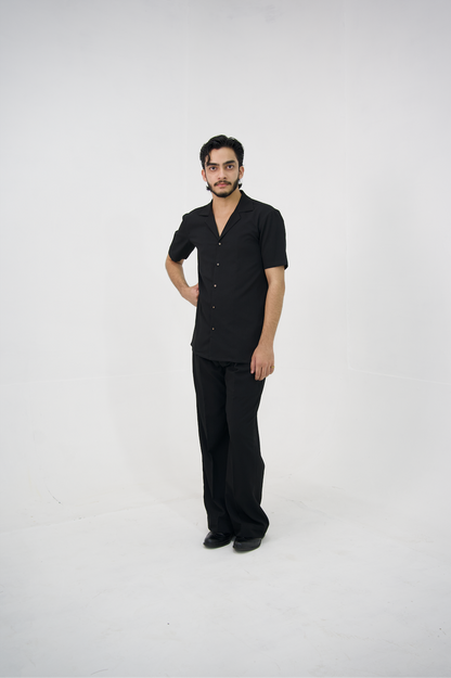 Man posing in a black cuban collar shirt and matching tailored trousers by Abbod Atelier on a white studio background.
